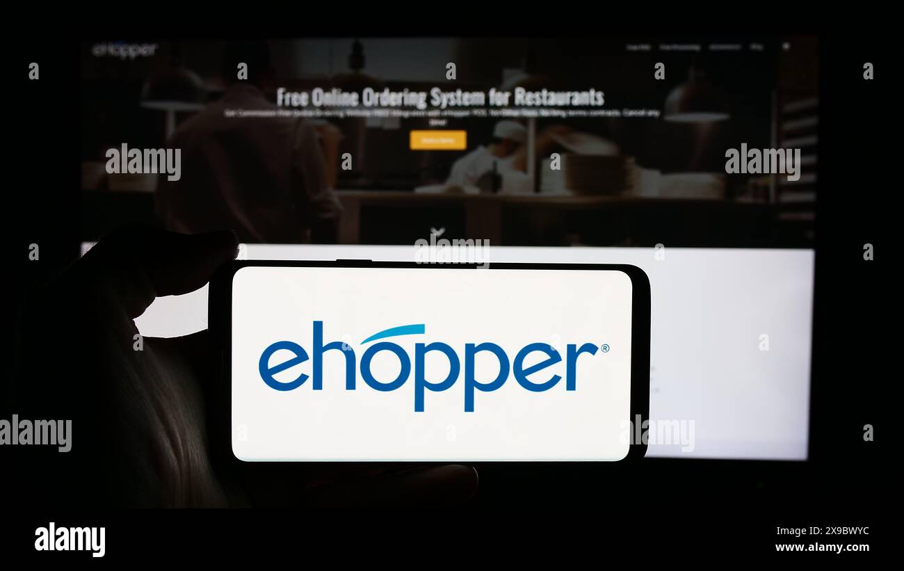 Person holding cellphone with logo of American point of sale systems company eHopper Services Inc. in front of webpage. Focus on phone display. Stock Photo