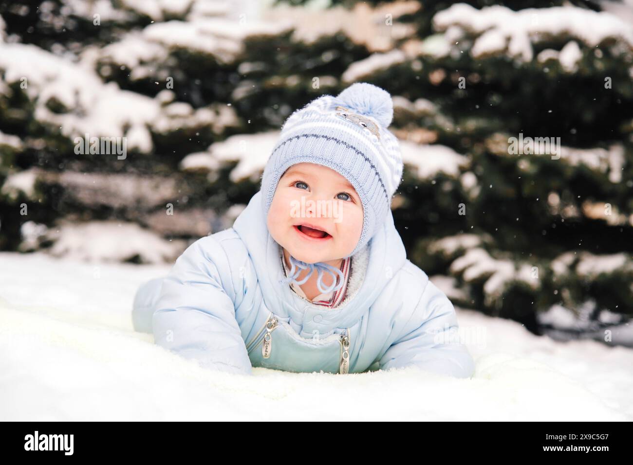 A baby in a blue jacket and knit hat enjoying the snow outdoors with a backdrop of snowy trees Stock Photo