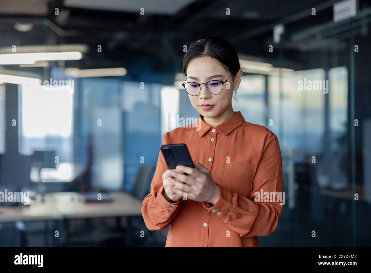 Asian woman using phone, serious thoughtful business woman inside office typing text message and browsing social media, female programmer software developer testing app online. Stock Photo