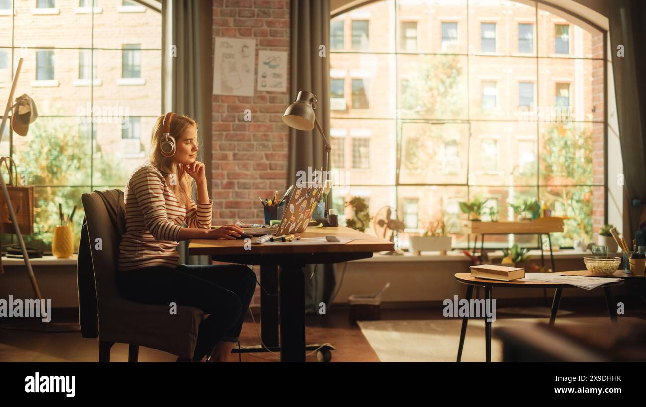 Young Pretty Woman with Blond Hair Wearing Headphones and Working from Home on Laptop Computer in Stylish Loft Apartment. Creative Female Smiling, Checking Social Media, Browsing Internet. Stock Photo