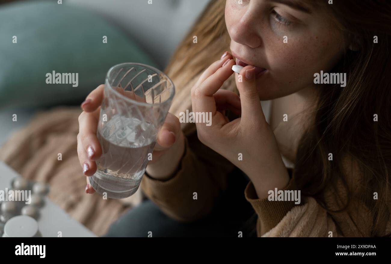 Caucasian young unhealthy woman sitting on sofa in the living room, covered with a blanket, holding a pill and a glass of water. Stock Photo