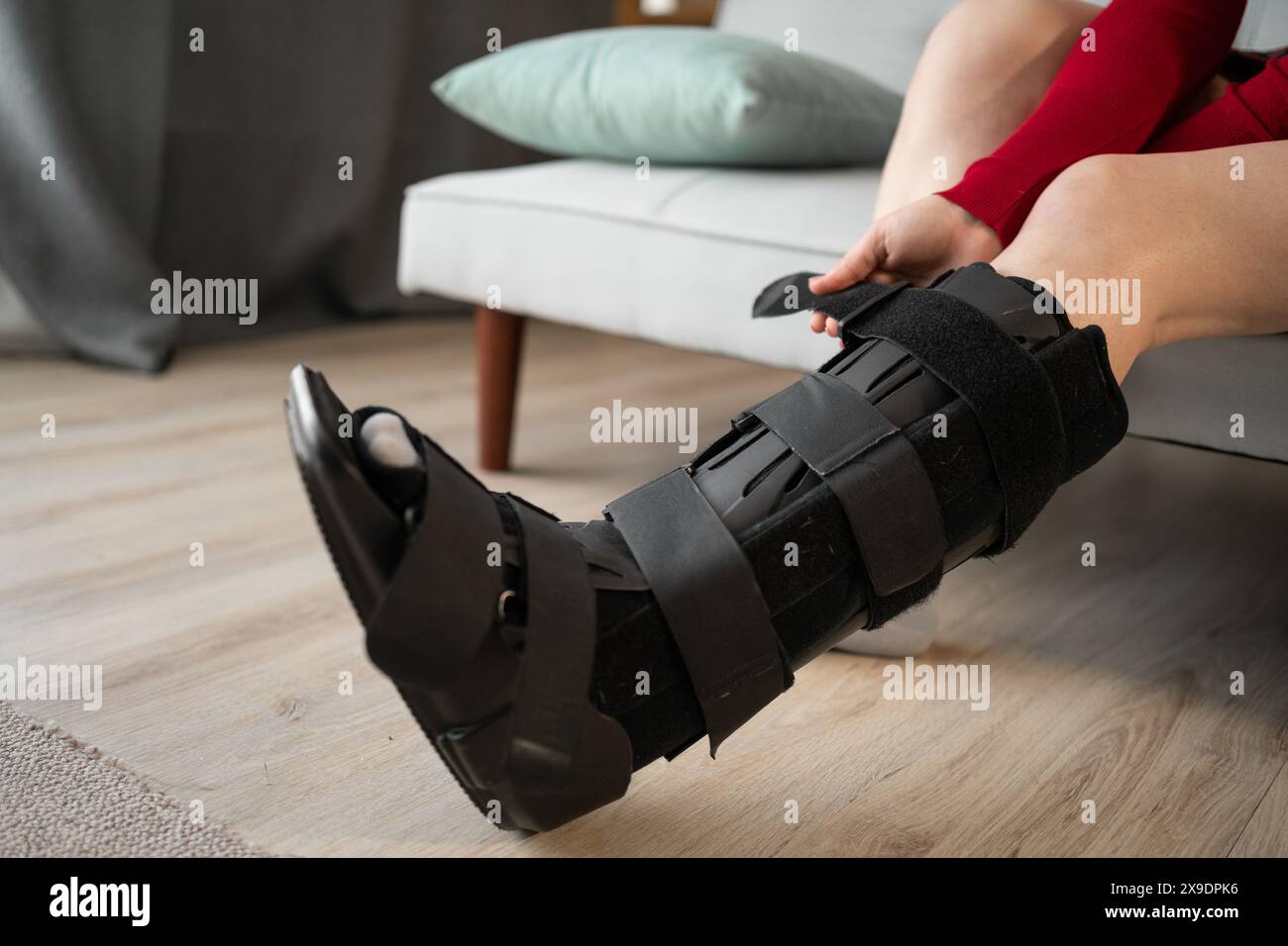 woman in orthosis sits on sofa after car accident. Attaching orthosis or bandage to support the ankle joint after leg surgery. Close-up. Copy space Stock Photo