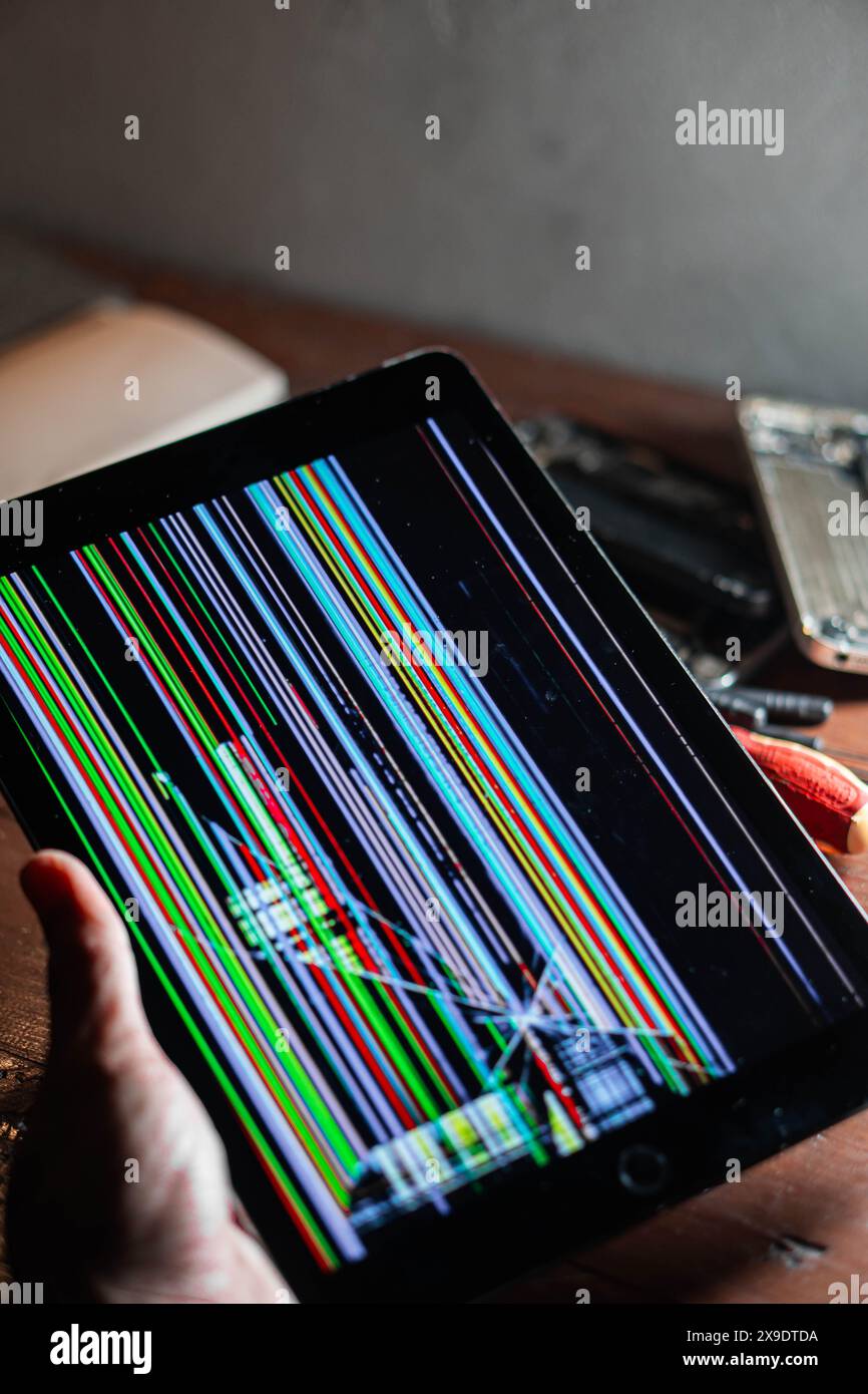 A person holding a tablet with a broken screen displaying colorful vertical lines. The background includes a wooden surface and some electronic tools. Stock Photo