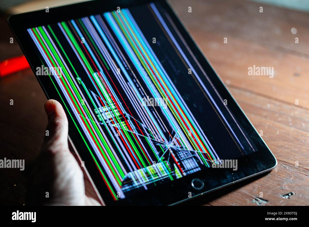 A person holding a tablet with a broken screen displaying colorful vertical lines and distorted images. The tablet is placed on a wooden surface. Stock Photo