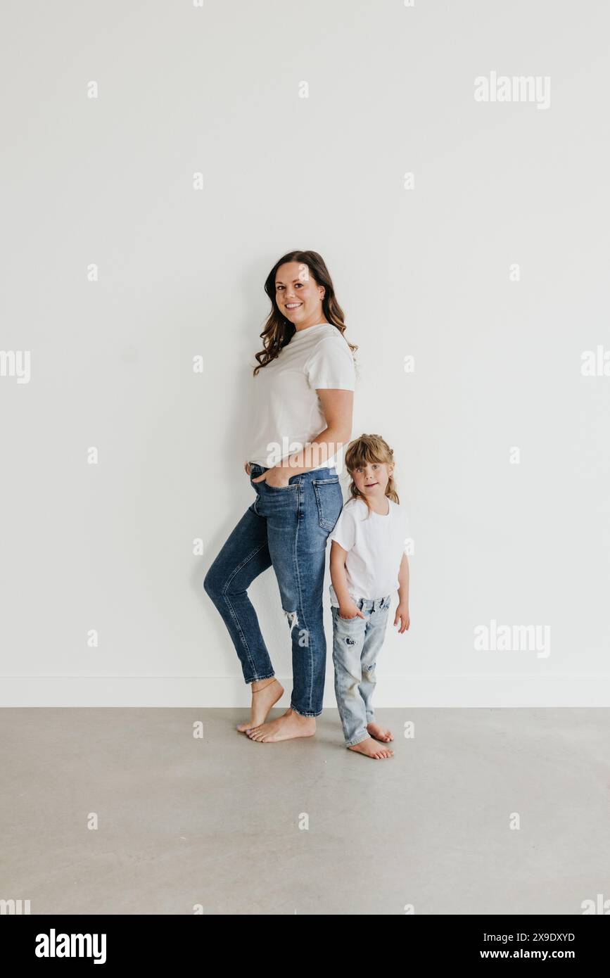 Mother and daughter stand back to back against white wall Stock Photo