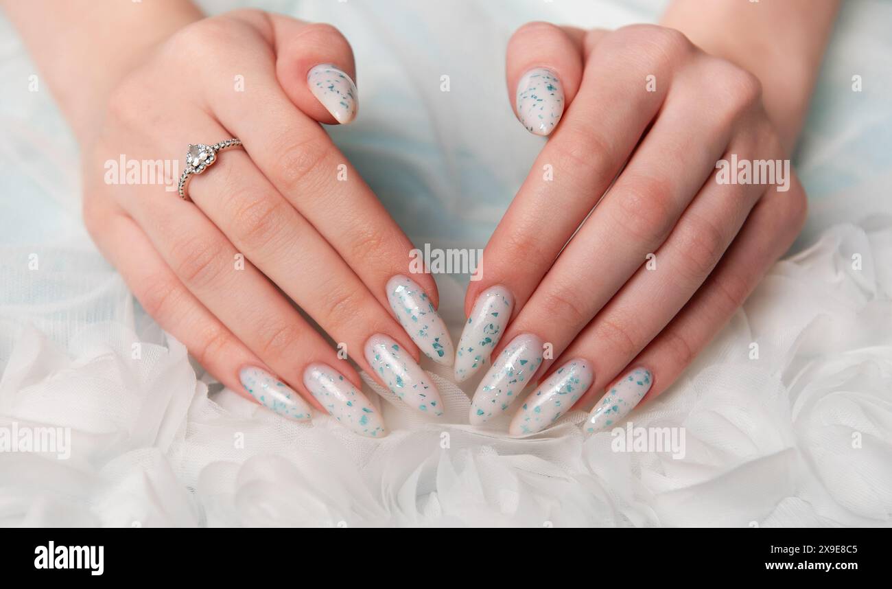 A close-up view of a persons elegantly manicured hands resting on soft, white fabric. The design features intricate blue floral art accented with glit Stock Photo