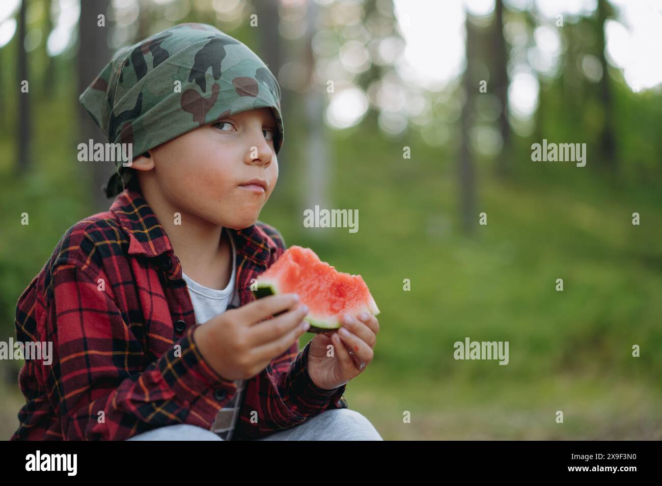 cute little caucasian boy wearing military bandana and plaid shirt eating watermelet in forest. Hiking, camping concept Stock Photo