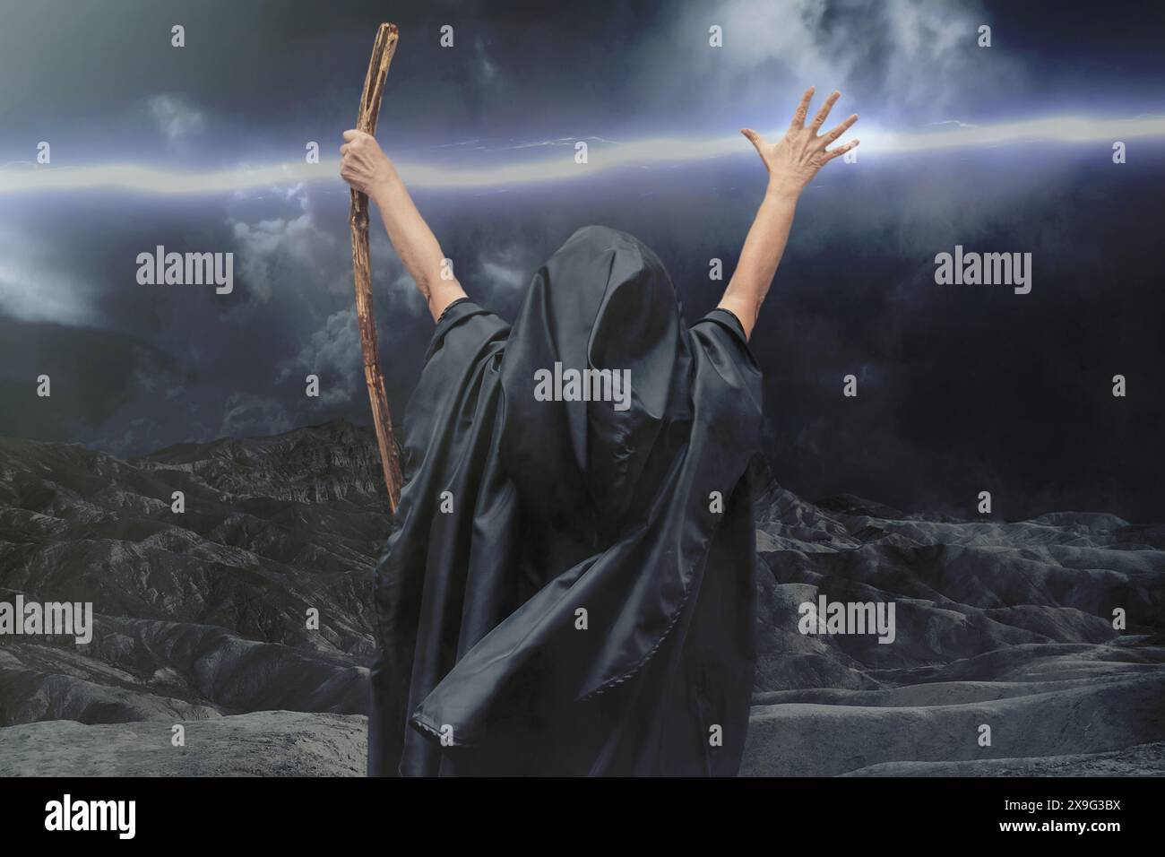 A sorcerer in a black hoodie stretched his arms with a staff up against the background of a stormy sky and lightning. Mysticism and magic, sorcerers a Stock Photo