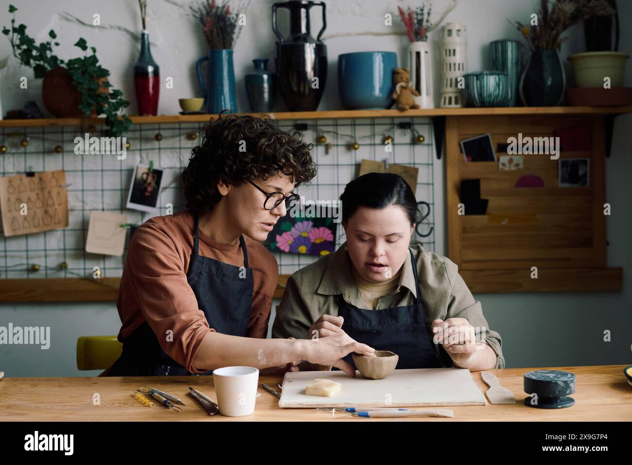 Mature female potter sitting at table teaching young woman with Down syndrome how to make ceramic vessel, copy space Stock Photo