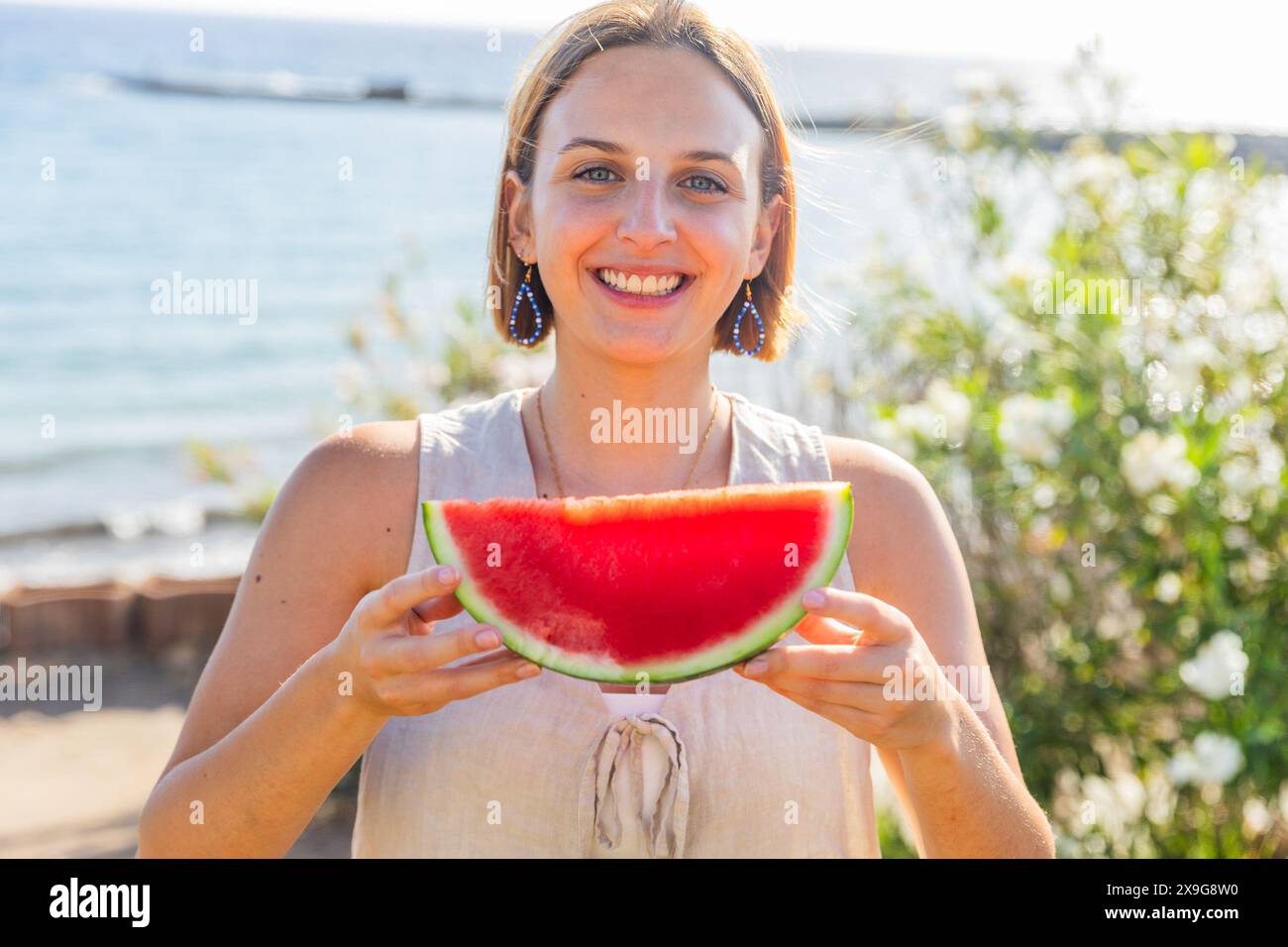 A woman is holding a watermelon slice in her hand, smiling at the camera. Stock Photo