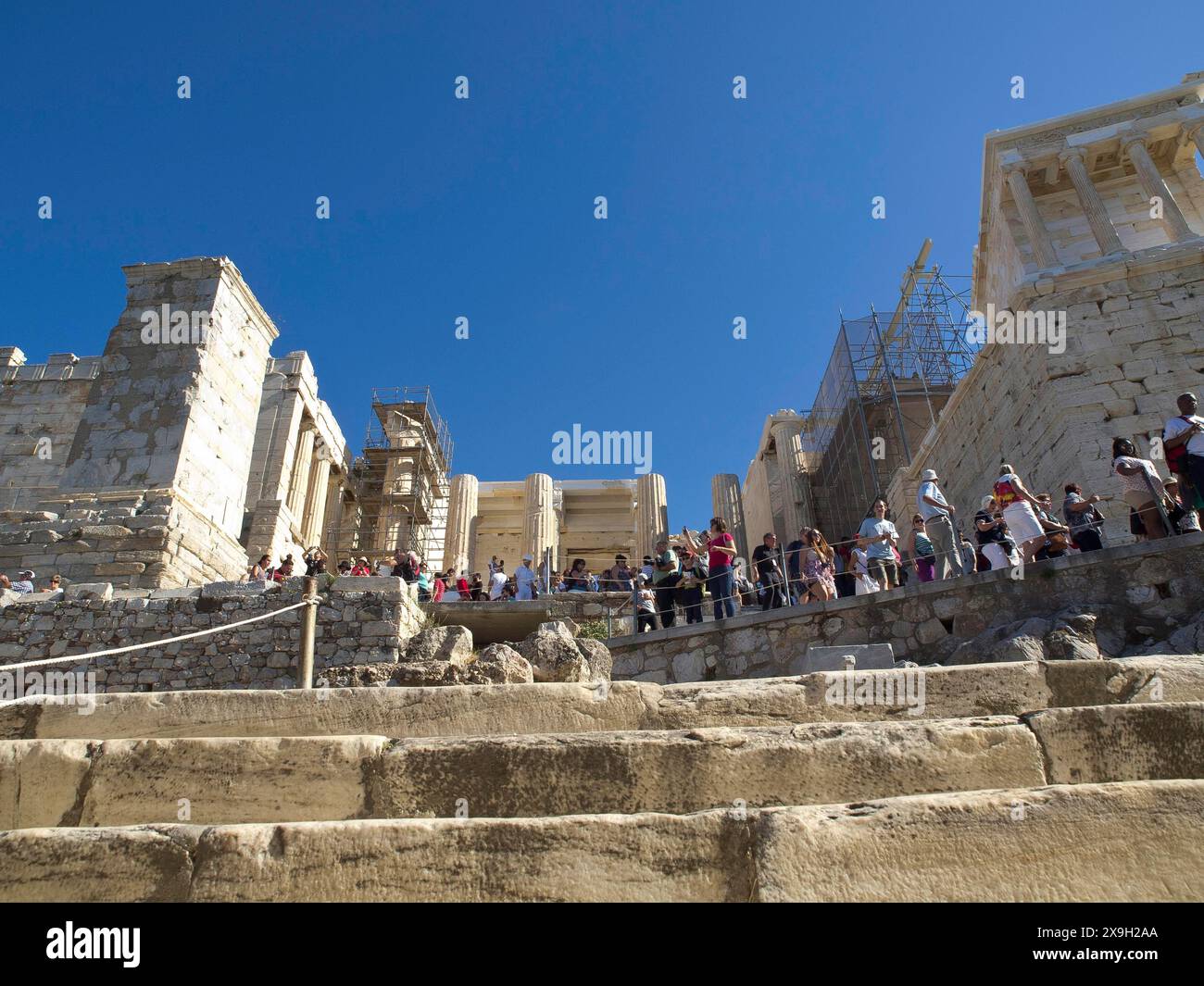 Many tourists climbing up the stone steps of a ruin on the Acropolis, Ancient buildings with columns and trees on the Acropolis in Athens against a Stock Photo