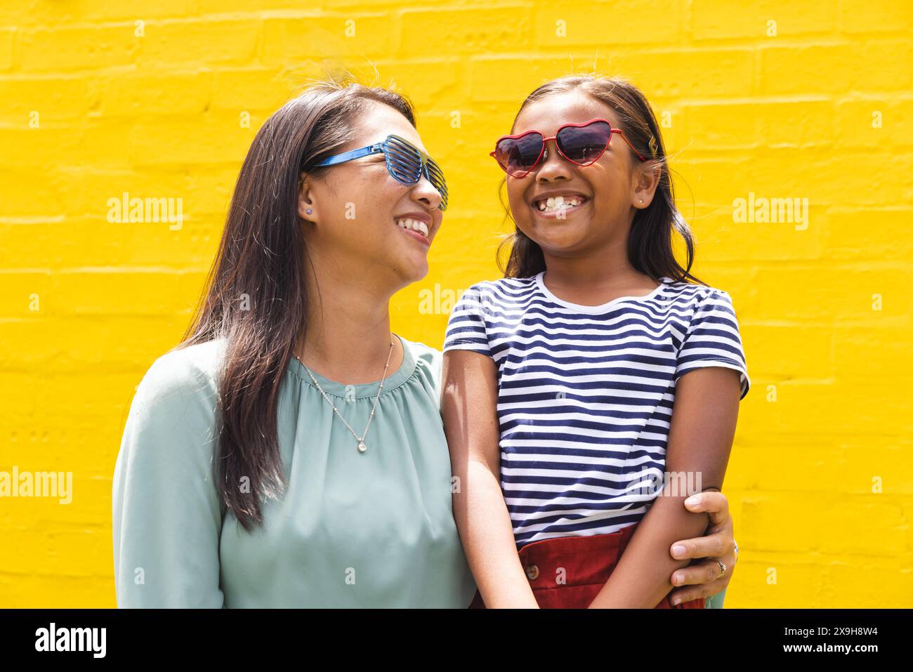 In front of a bright yellow wall outdoors, a biracial young girl and a woman are smiling Stock Photo