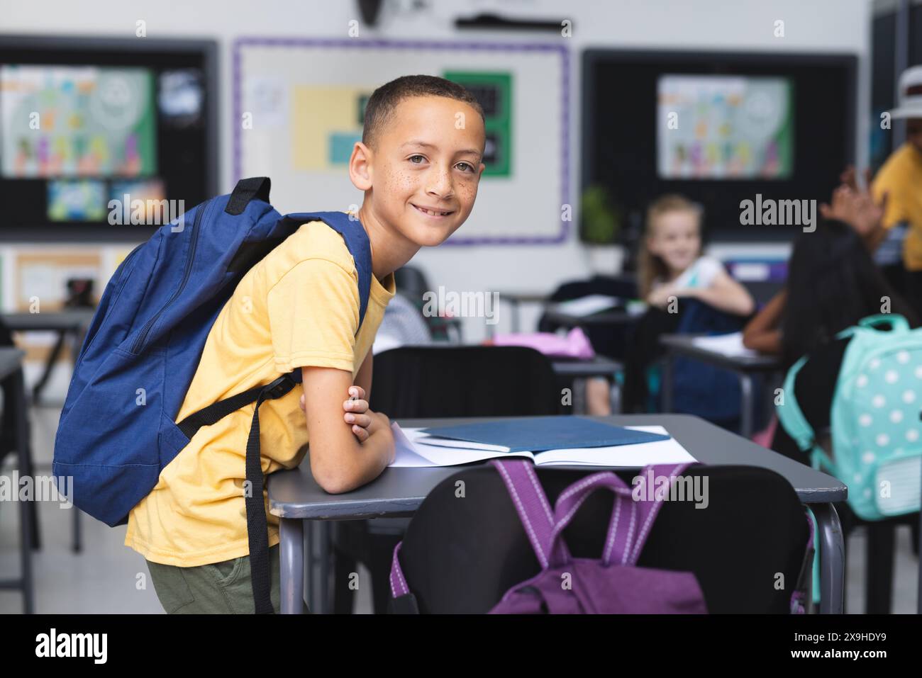 Biracial boy with a backpack stands in a school classroom, smiling ...