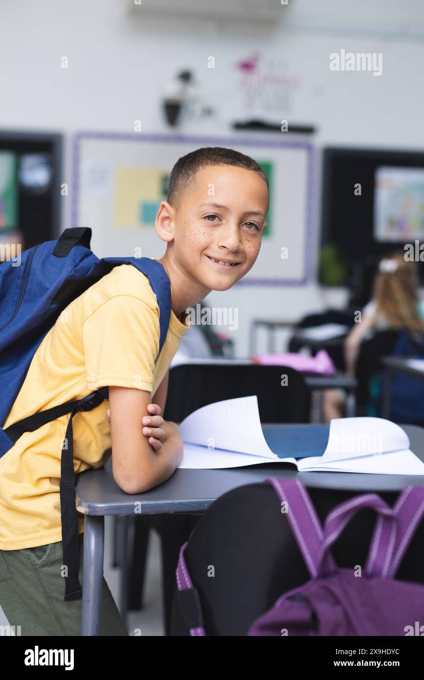 Biracial boy with a backpack smiles at a desk in a school classroom ...