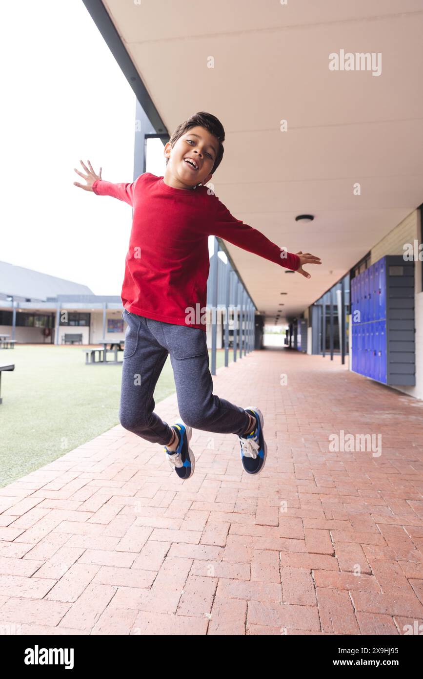 In school outdoors, biracial young boy wearing a red shirt is jumping ...