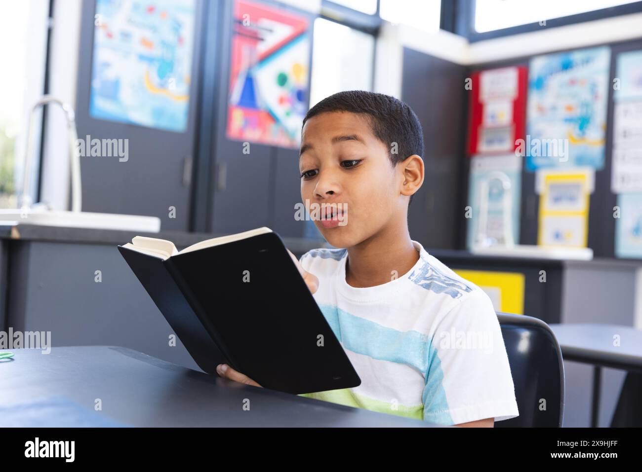 Biracial boy focuses on reading in a classroom adorned with educational ...