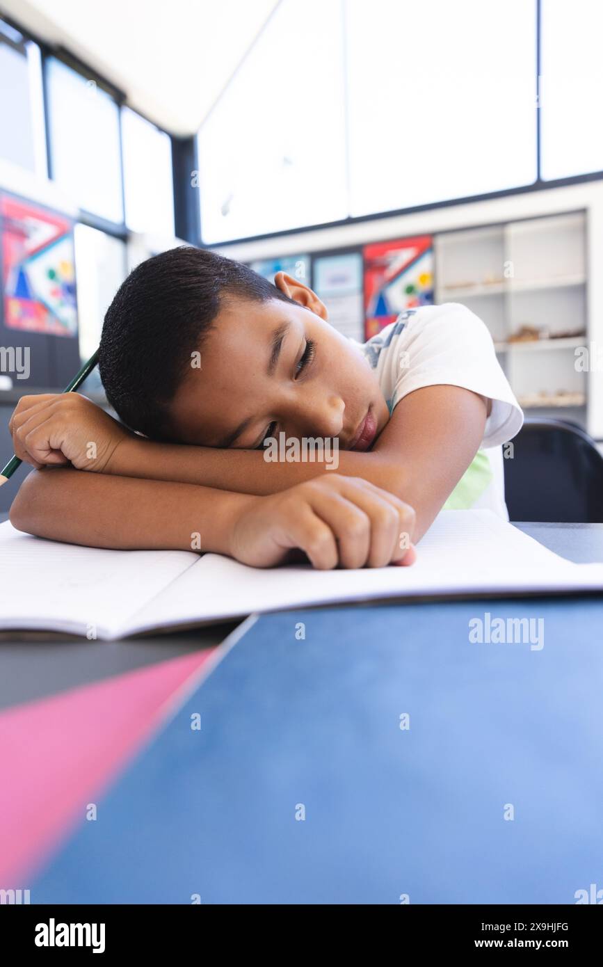In a school setting, a young biracial student rests his head on his arms with copy space. Surrounded by chemistry signs, he appears tired or thoughtfu Stock Photo
