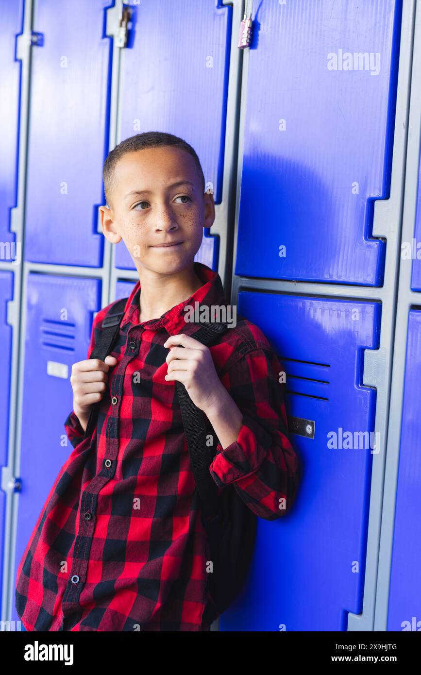 Biracial boy with a backpack stands by blue lockers in school. He wears ...