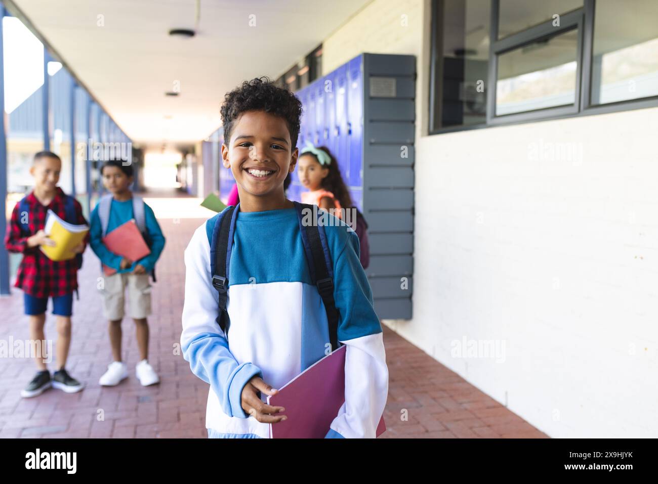 Biracial boy with a bright smile stands in a school hallway, holding a folder. Behind him, two biracial boys and two biracial girls carry backpacks, s Stock Photo