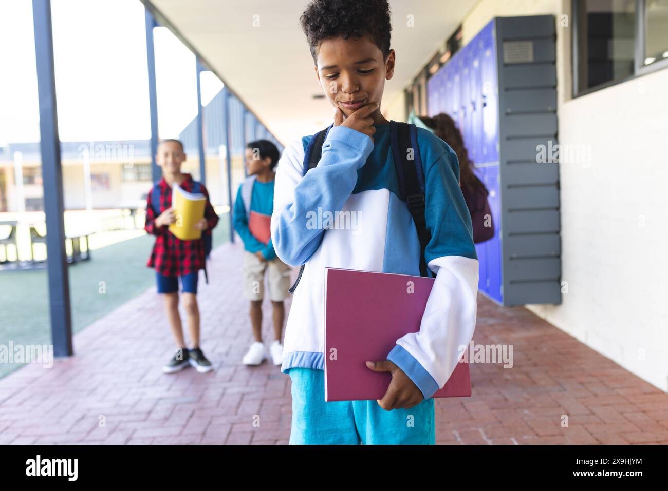 Biracial boy in blue stands thoughtfully in a school corridor, holding ...