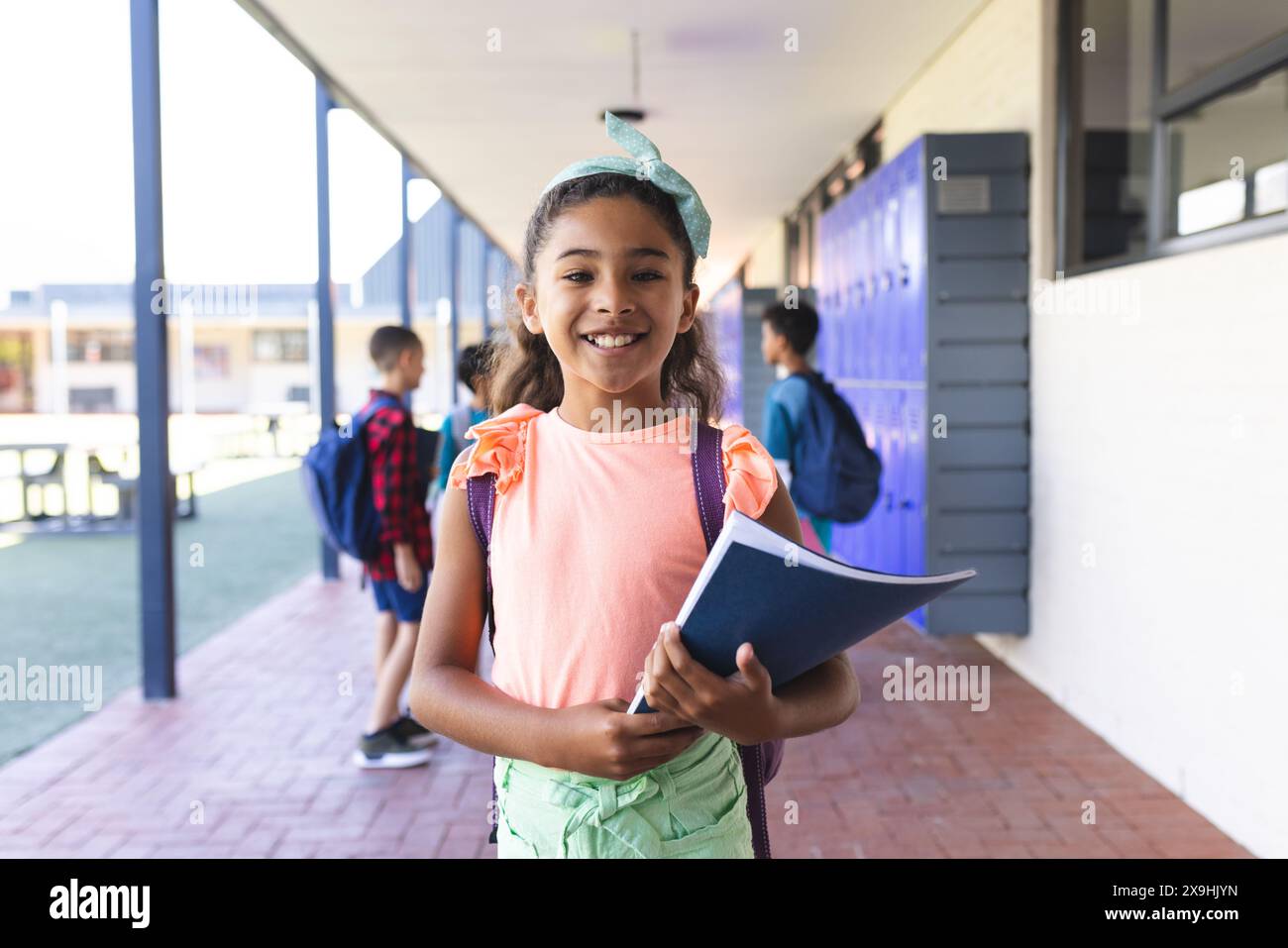 Biracial girl with a blue book stands smiling in a school corridor Stock Photo
