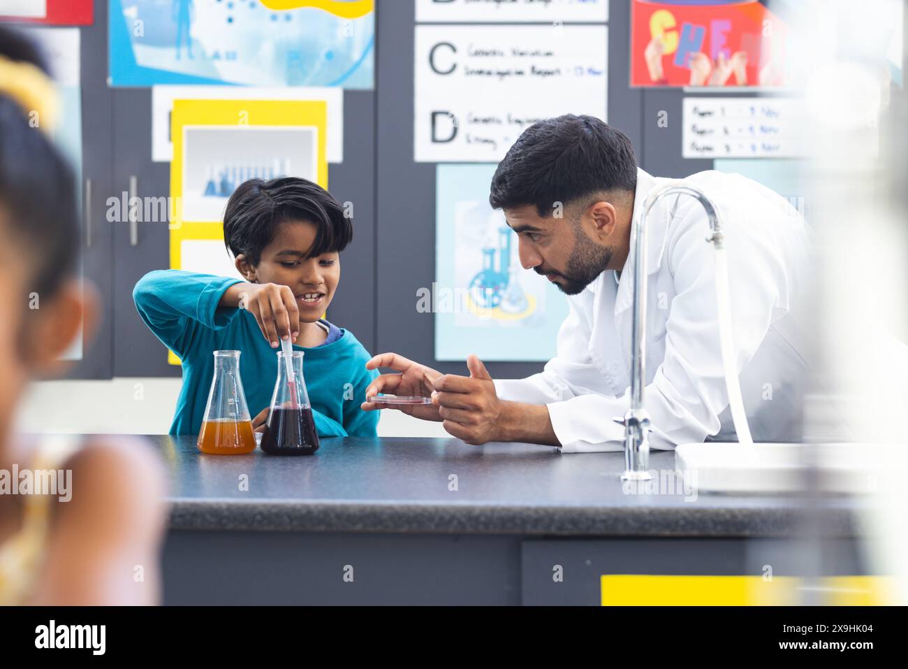 In school, Asian male teacher guiding biracial male student in doing a science experiment. Both are examining colorful liquids in flasks, surrounded b Stock Photo