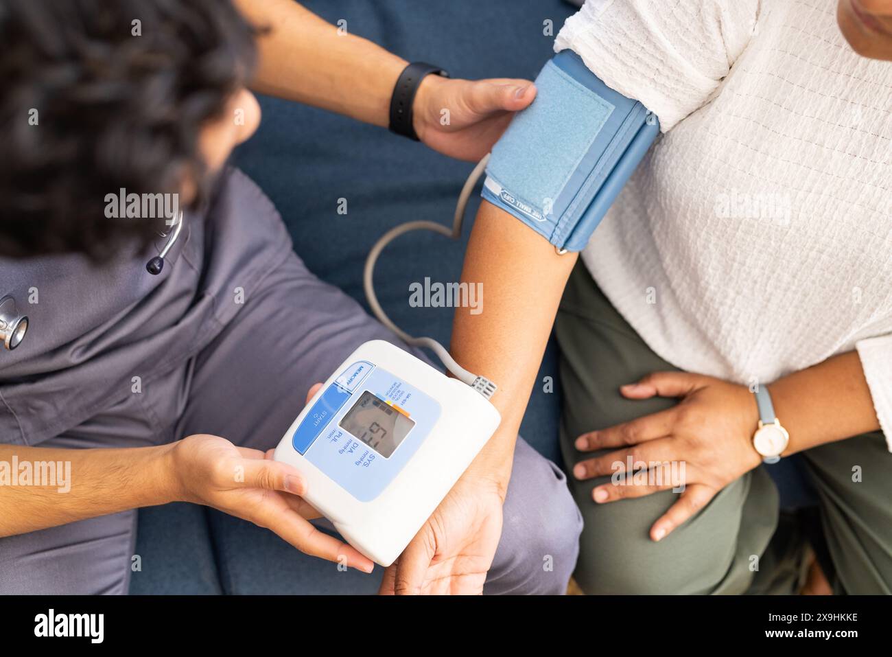 At home, a young biracial male nurse checks an elderly woman's blood pressure. They are in a medical office with a blue background and medical equipme Stock Photo