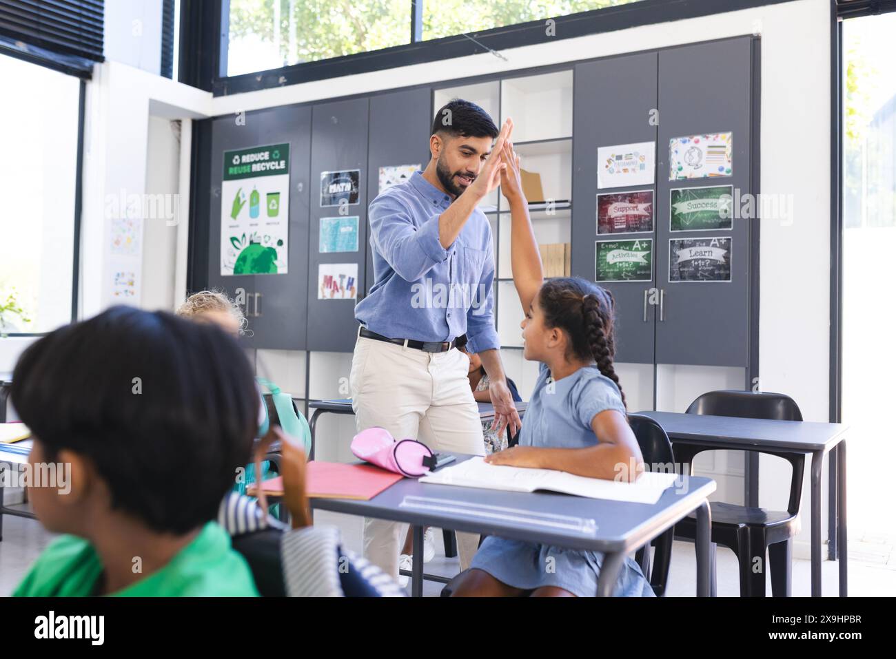 Young Asian male teacher high-fives a biracial girl in a school classroom Stock Photo