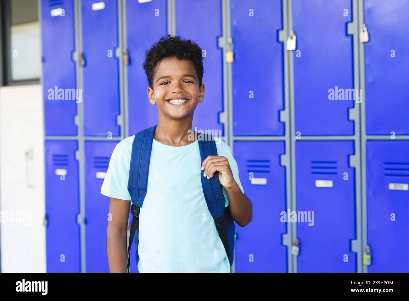 Biracial boy with a backpack stands smiling in front of blue lockers in ...