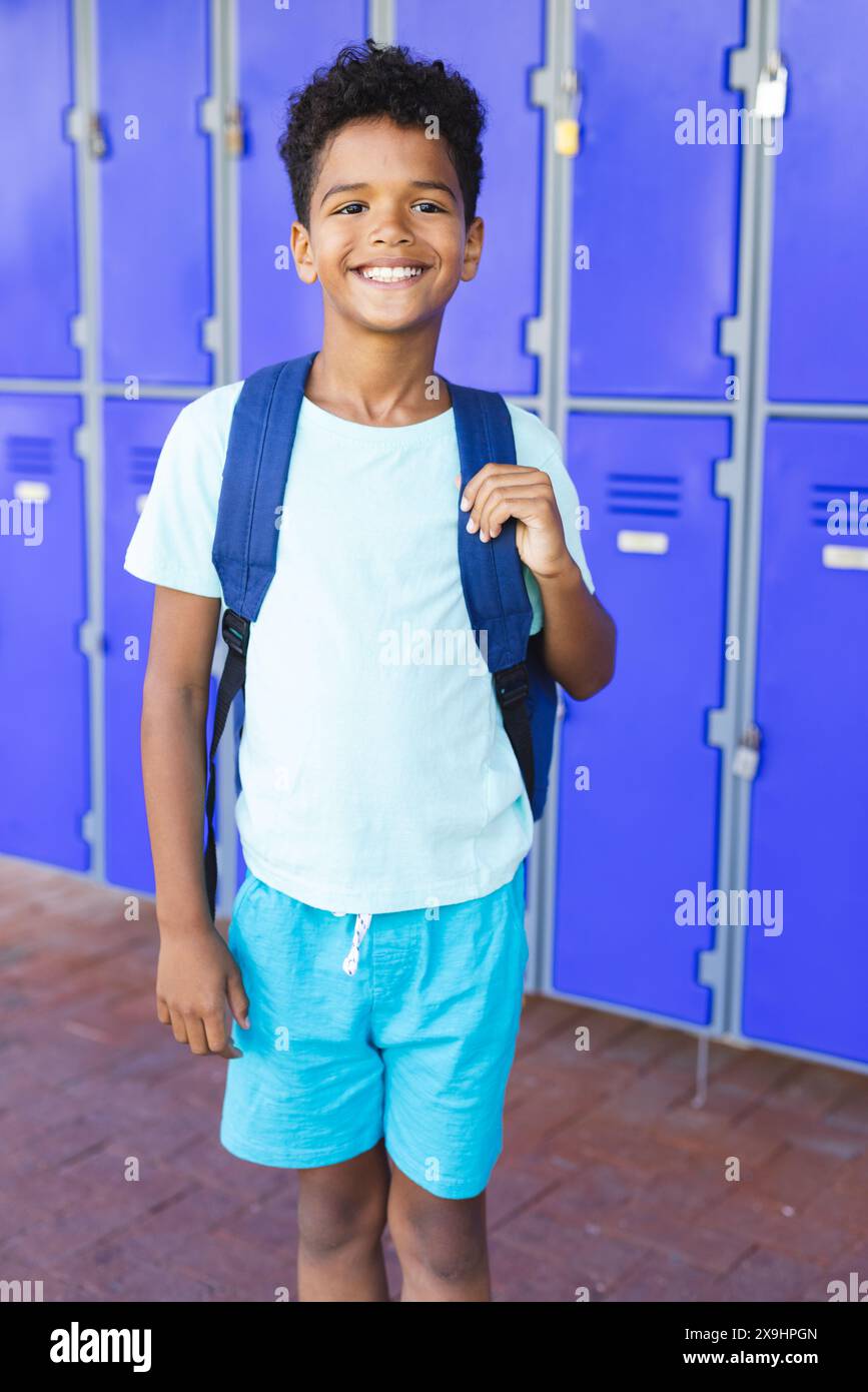 A biracial boy with a bright smile stands in front of blue lockers in ...