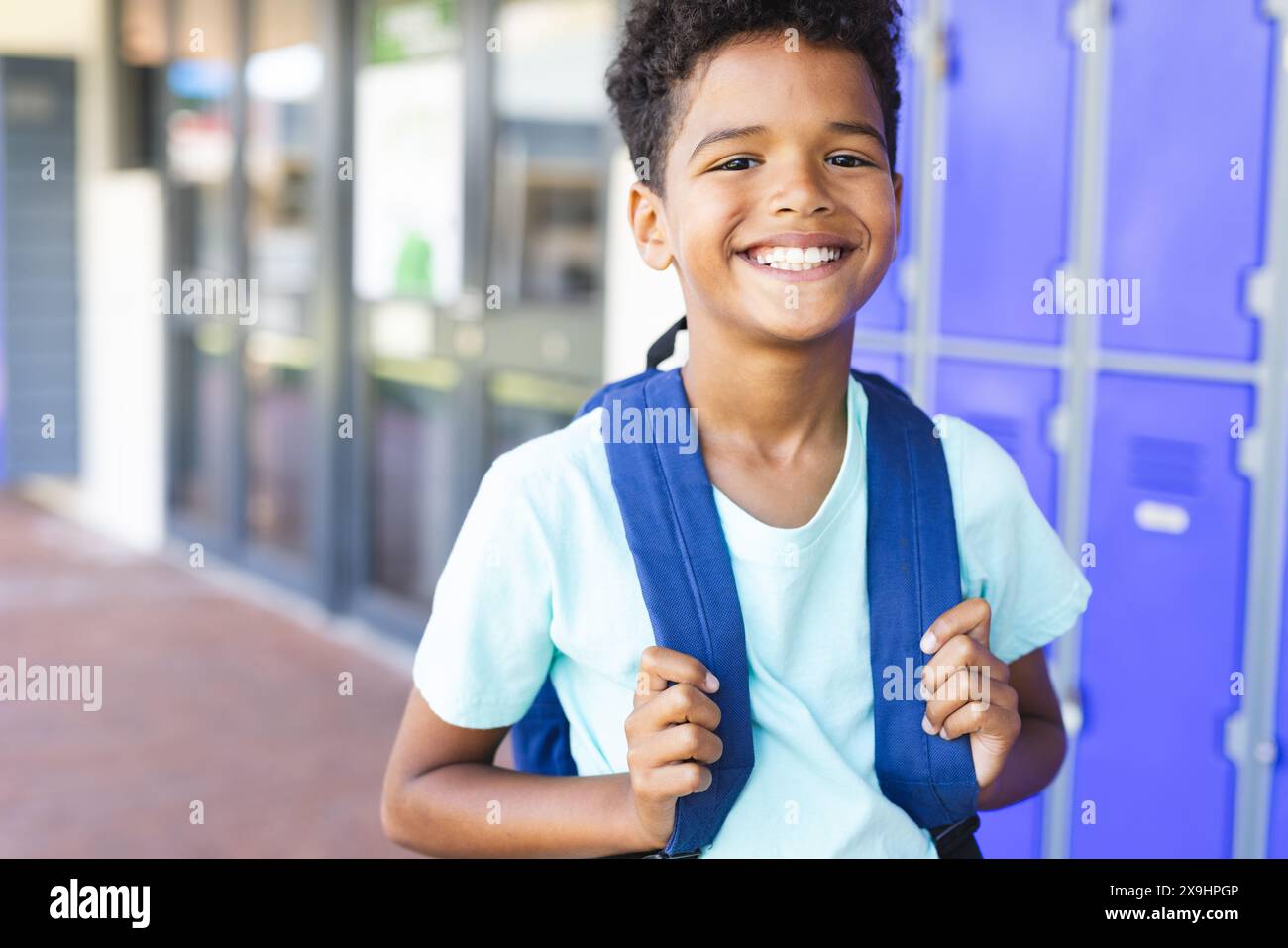 Biracial boy with a bright smile stands in front of lockers at school ...