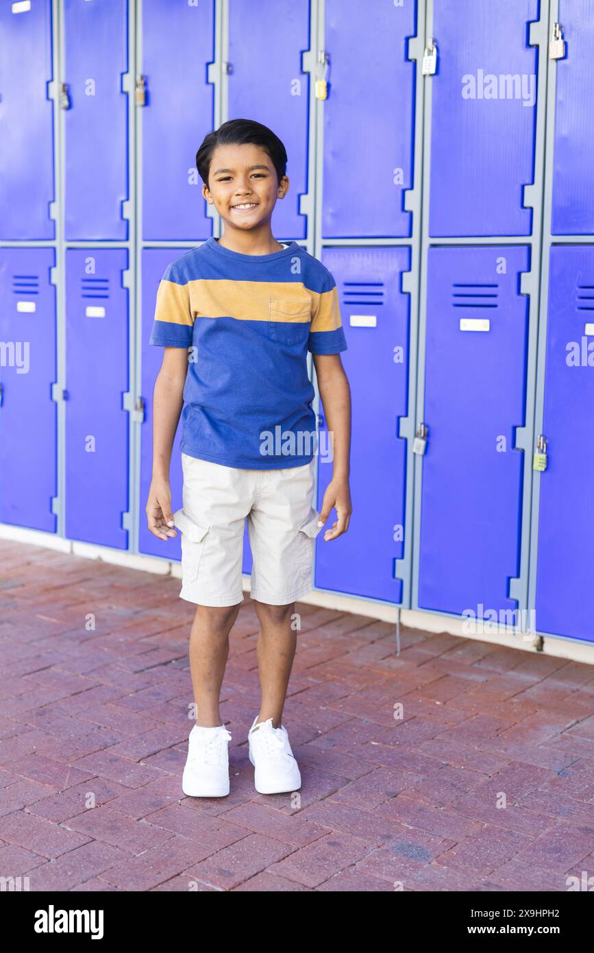 Biracial boy stands smiling in front of blue lockers at school, dressed ...