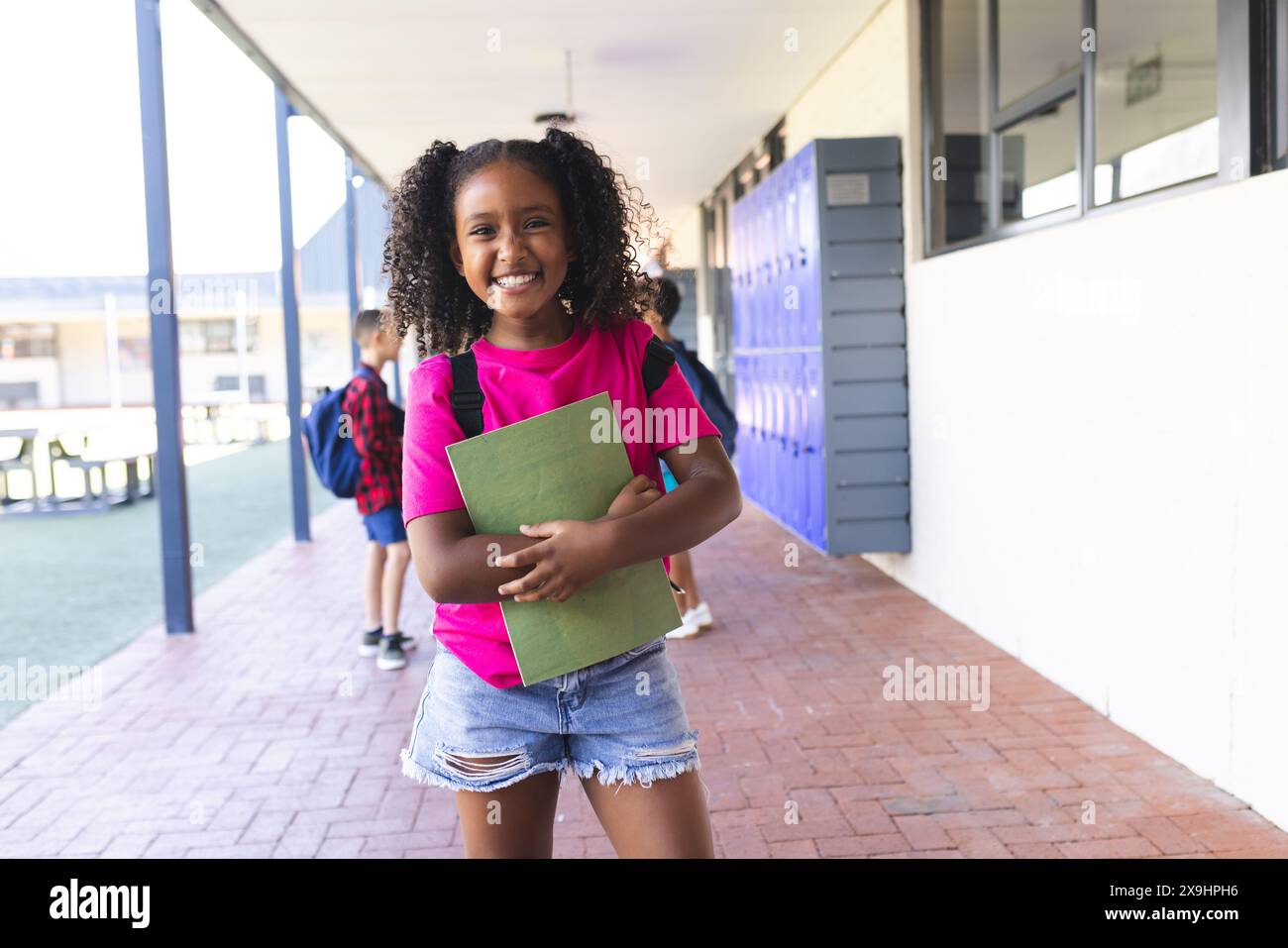Biracial girl with curly hair smiles, holding a green book at school Stock Photo