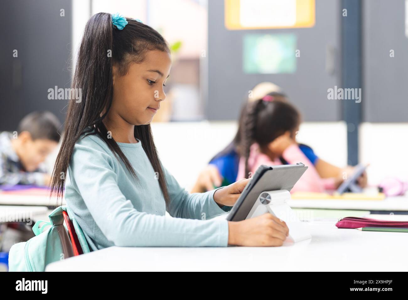 In school, young biracial girl holding tablet sits at a desk in ...