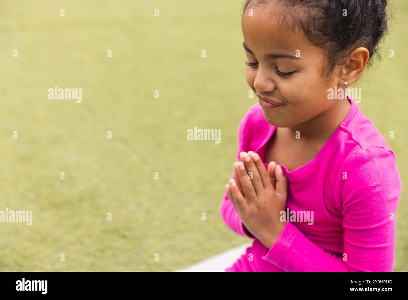 Young biracial girl in pink prays and does yoga on green grass at school with c Stock Photo