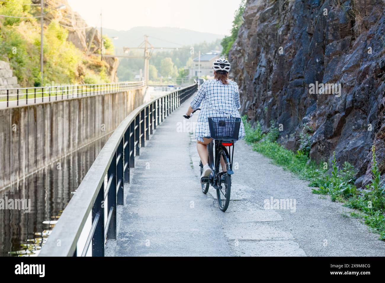 Mature woman in helmet cycling along a gateway channel. View from behind. Adult people active lifestyle concept. Stock Photo