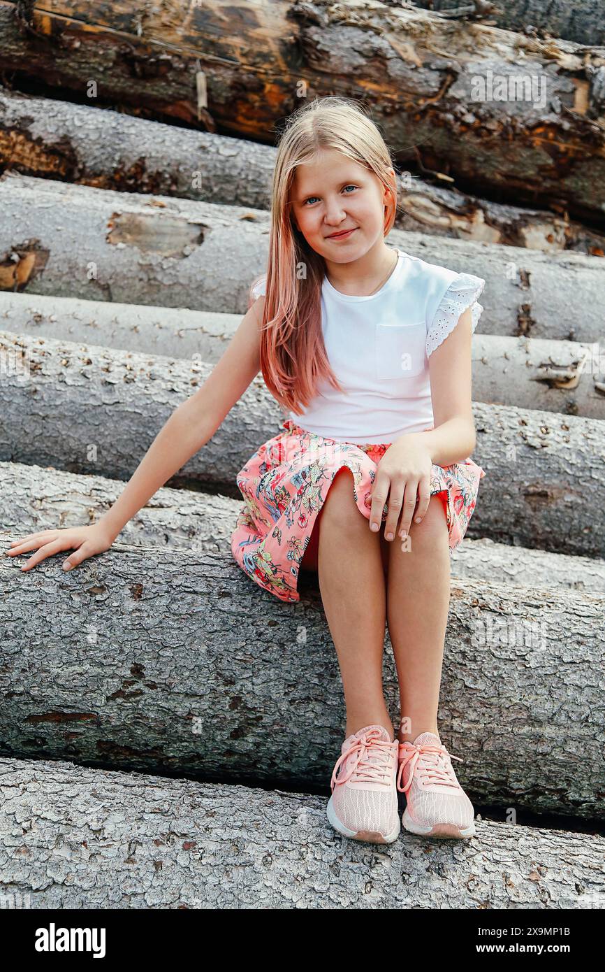 A young girl with pink-tipped hair sits on tree logs wearing a white shirt, floral skirt, and pink shoes, Belarus, Minsk Stock Photo