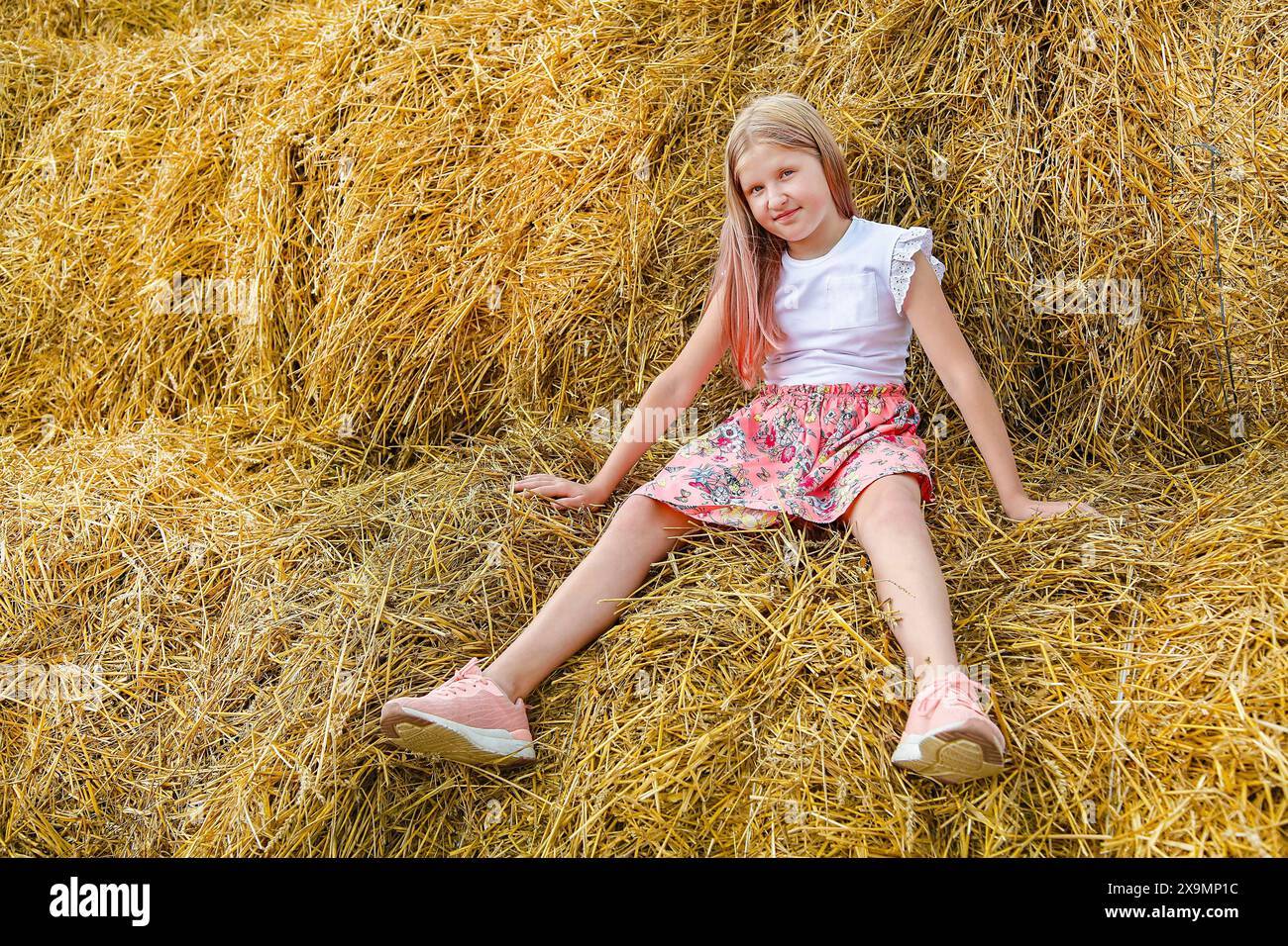 A young girl with blonde hair sits relaxed on a haystack, wearing a white shirt, floral skirt, and pink shoes against a rural countryside backdrop Stock Photo