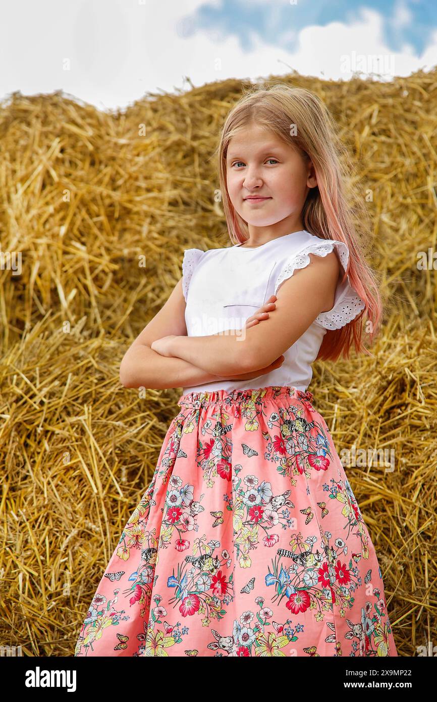 A young girl with crossed arms stands confidently in front of a haystack, wearing a floral skirt and white top against a summer sky, Belarus, Minsk Stock Photo