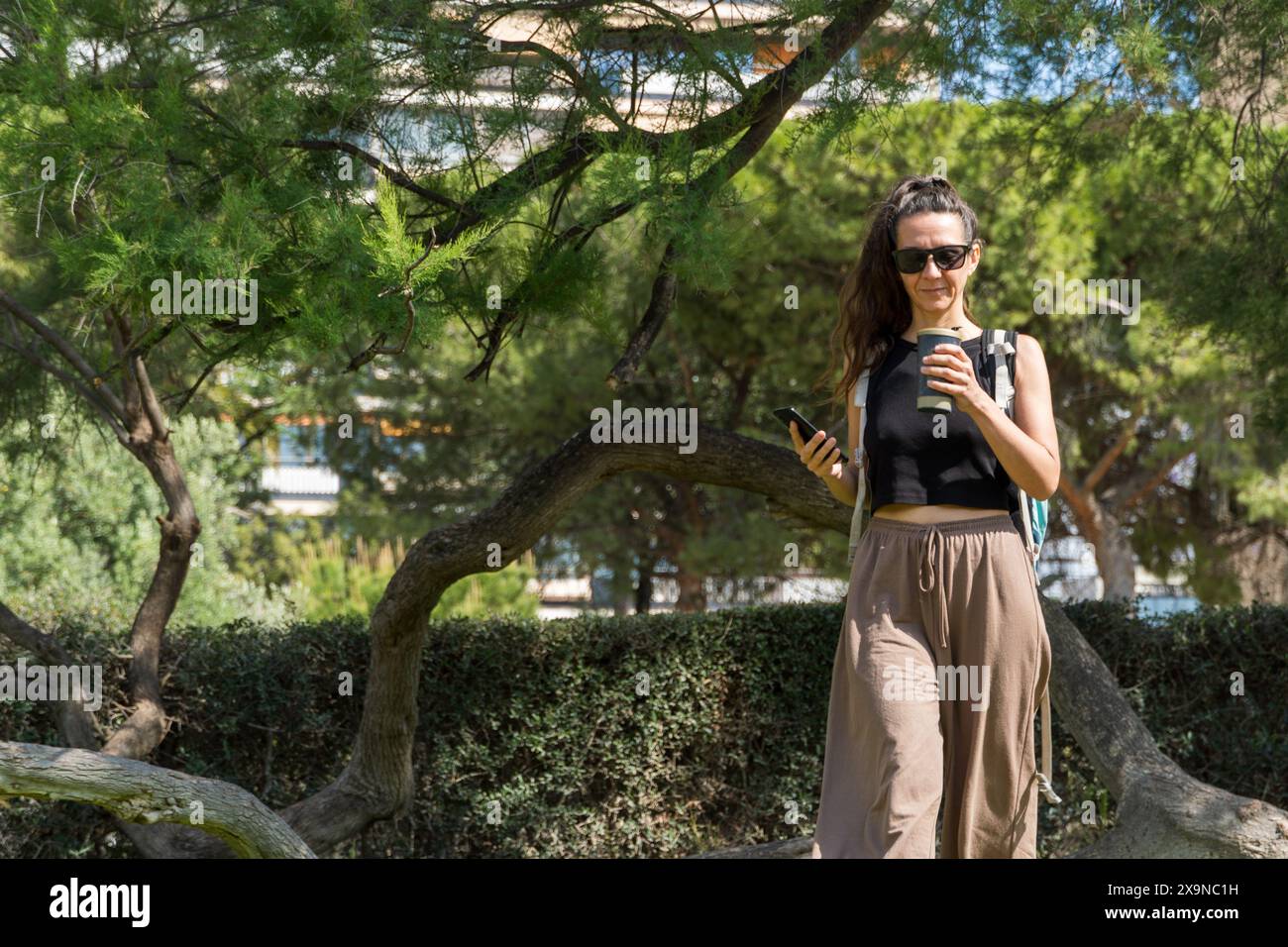 A young woman walks in a park, casually dressed, wearing sunglasses, holding a coffee cup and a smartphone Stock Photo