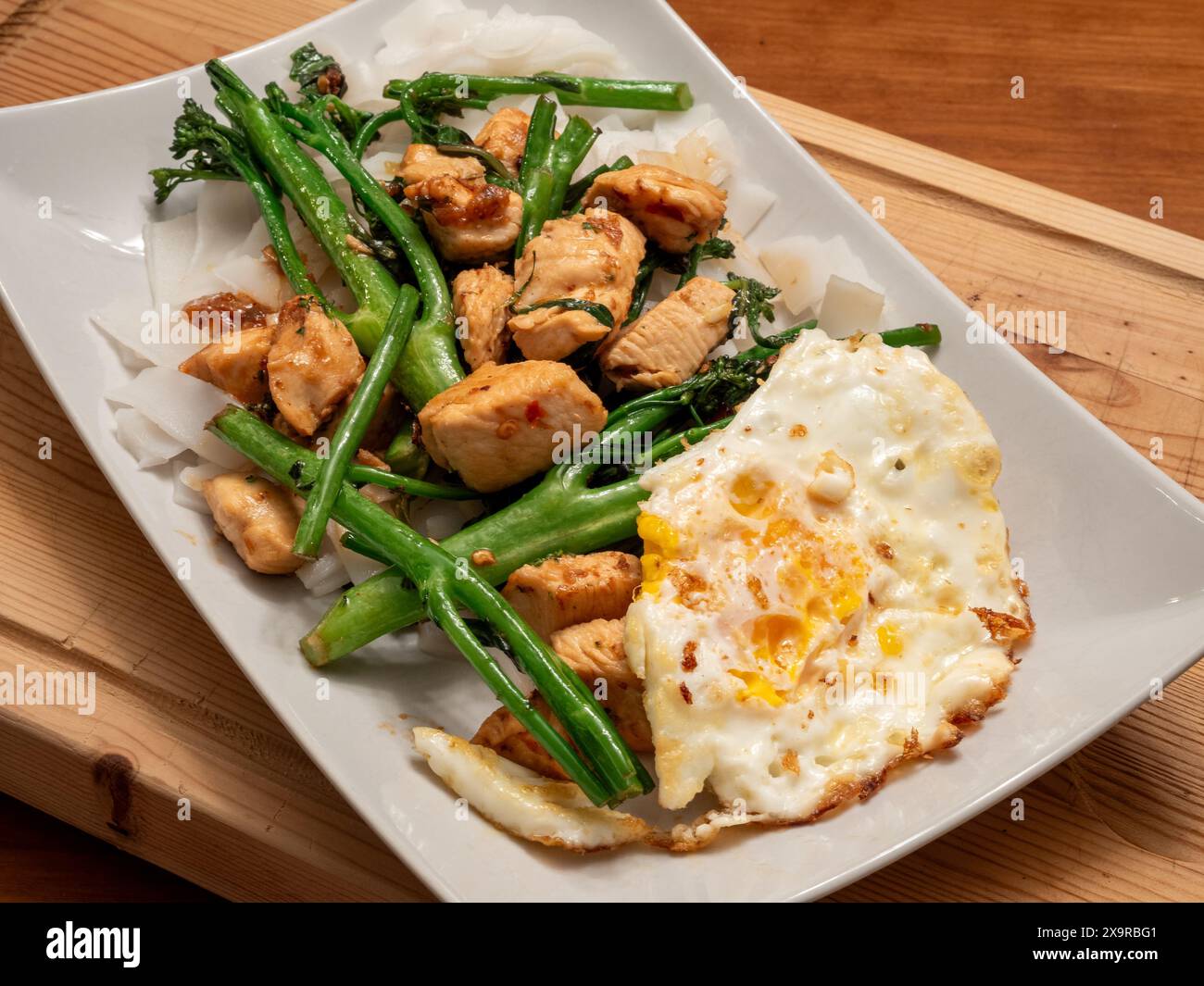 A close-up of a white plate overflowing with an Asian stir-fry: crispy chicken, vibrant broccolini, a perfectly fried egg, and wide, tempting noodles. Stock Photo