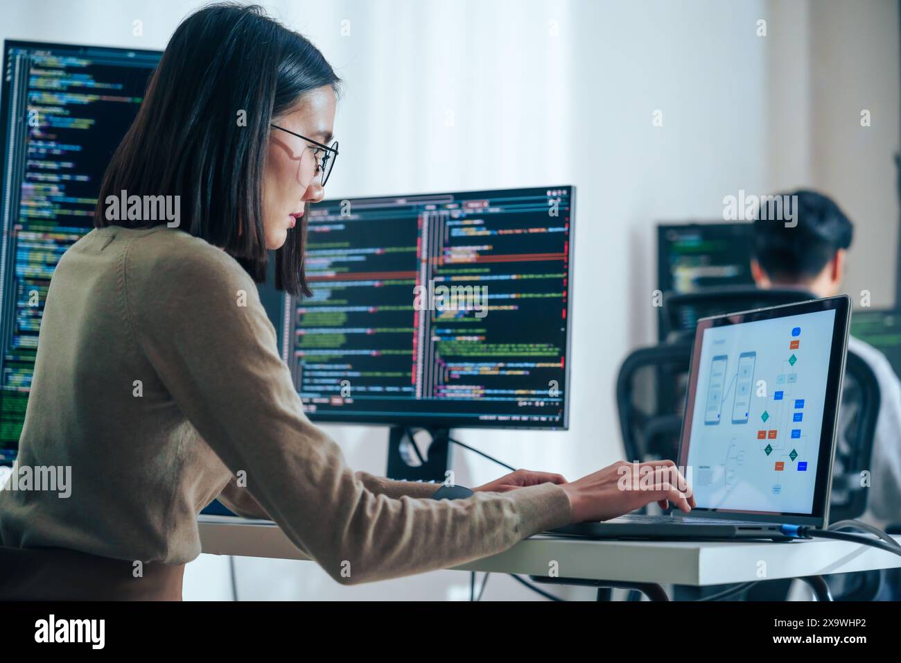 Asian software developers working on multiple screens displaying code and application diagrams in a modern office Stock Photo