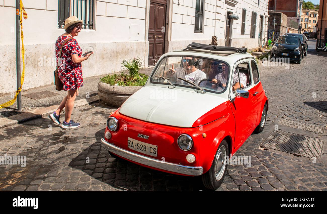 Three smiling people drive along the old Trastevere area of Rome in their vintage white and red Fiat 500 while a passer by looks and smiles. Stock Photo