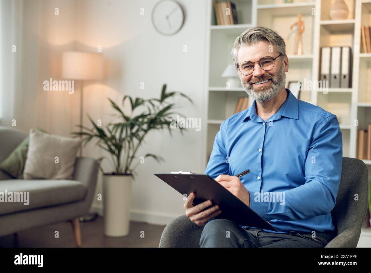 Gray haired man psychologist wearing blue shirt holding clipboard standing in his office Stock Photo