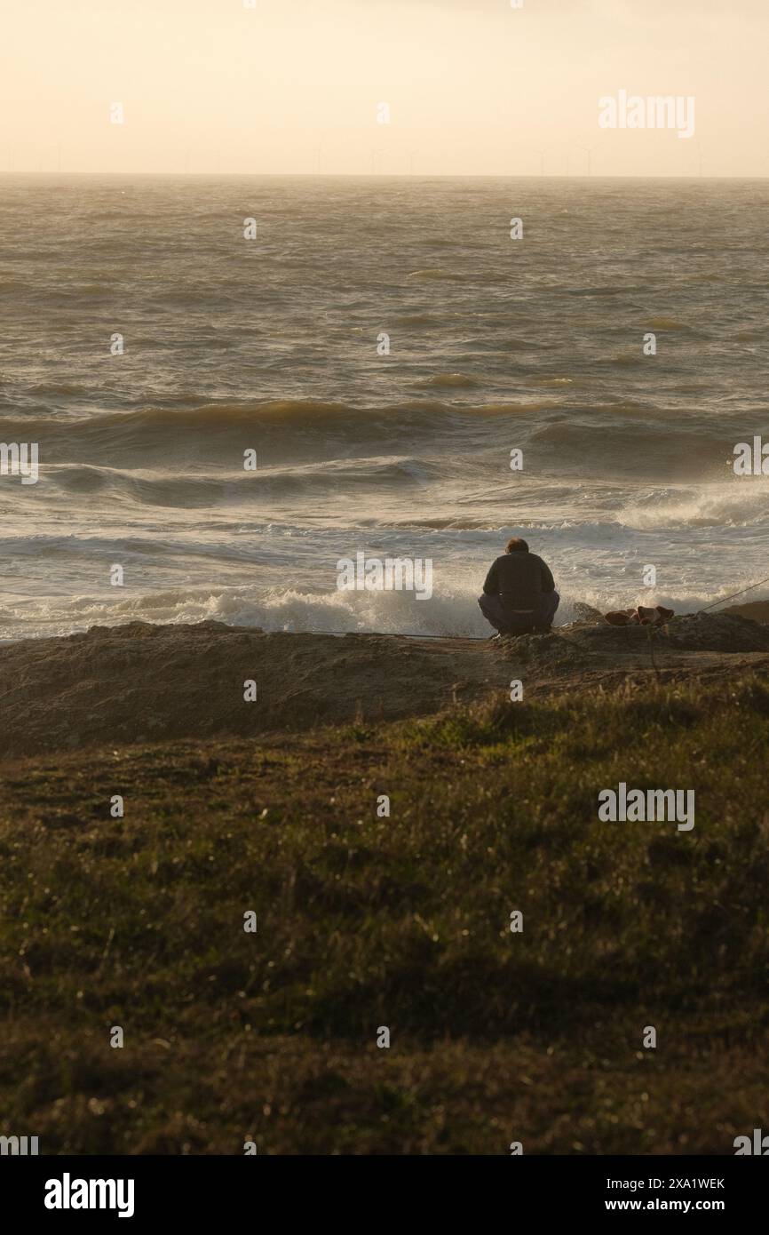 A man sitting on the shore, watching waves at sunset Stock Photo