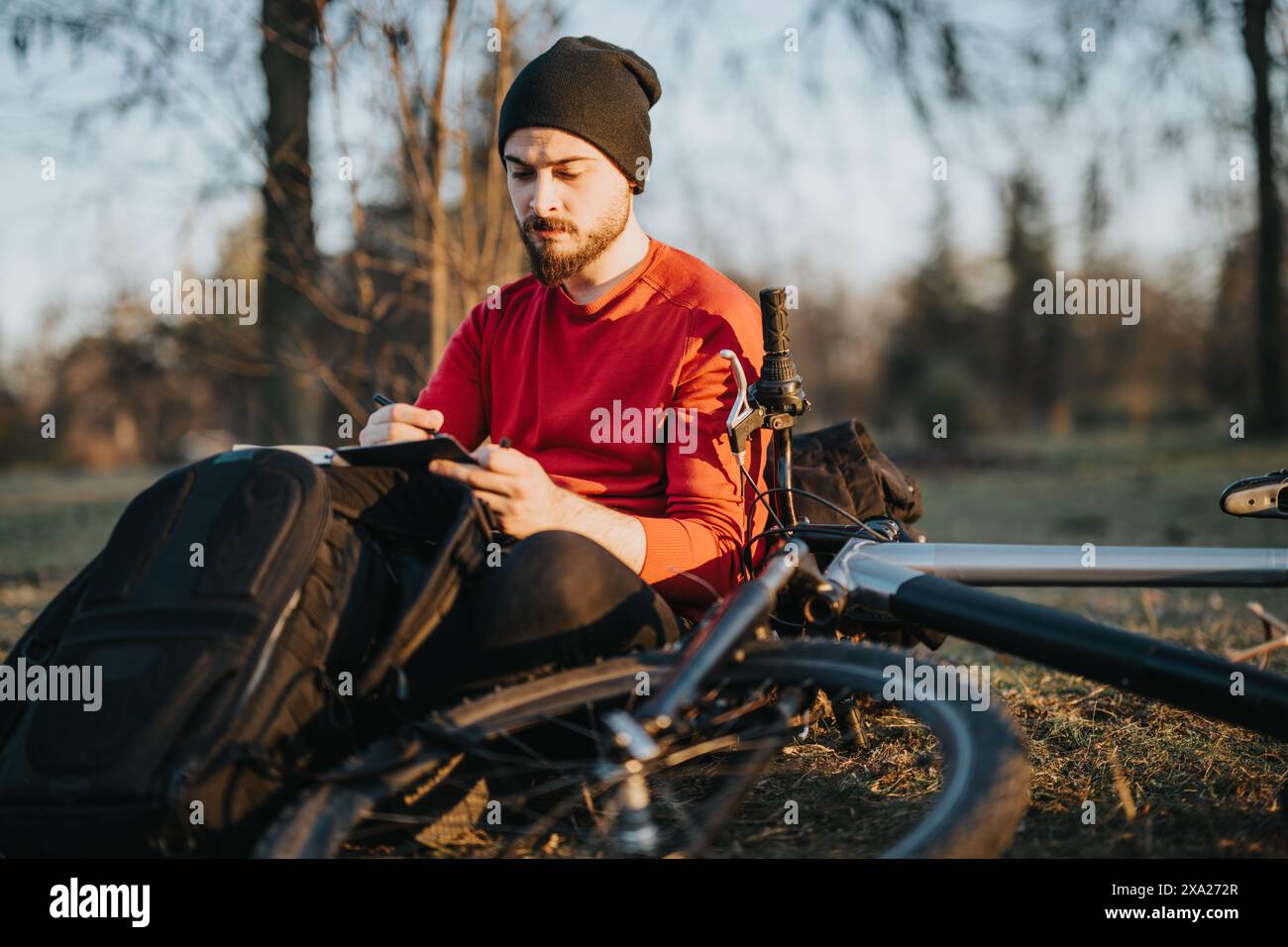 Young man pausing bike ride in park to write or draw in the notebook ...