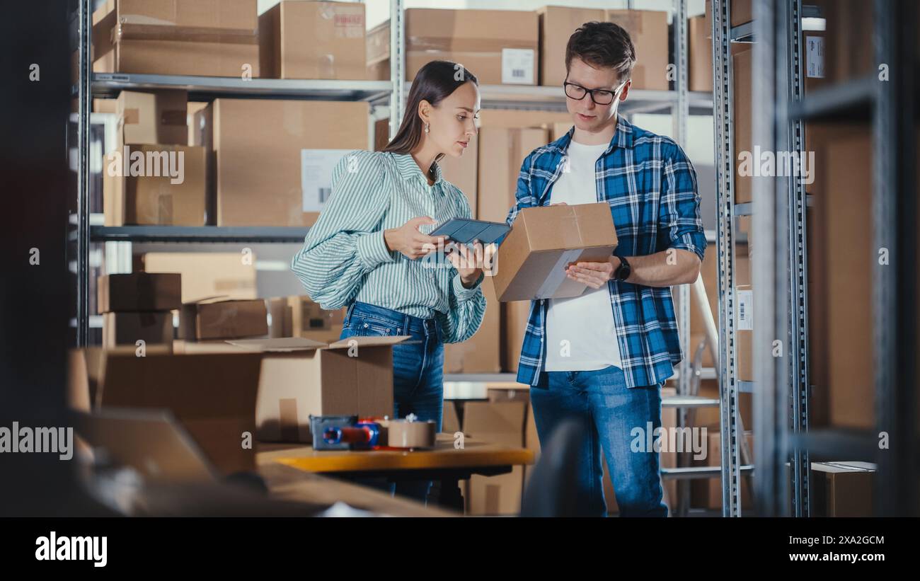 Female Sales Manager Using Tablet Computer, Talking to a Worker Holding a Cardboard Package, They Discuss Work and Smile. Warehouse with Parcels Ready for Shipment in the Background. Stock Photo