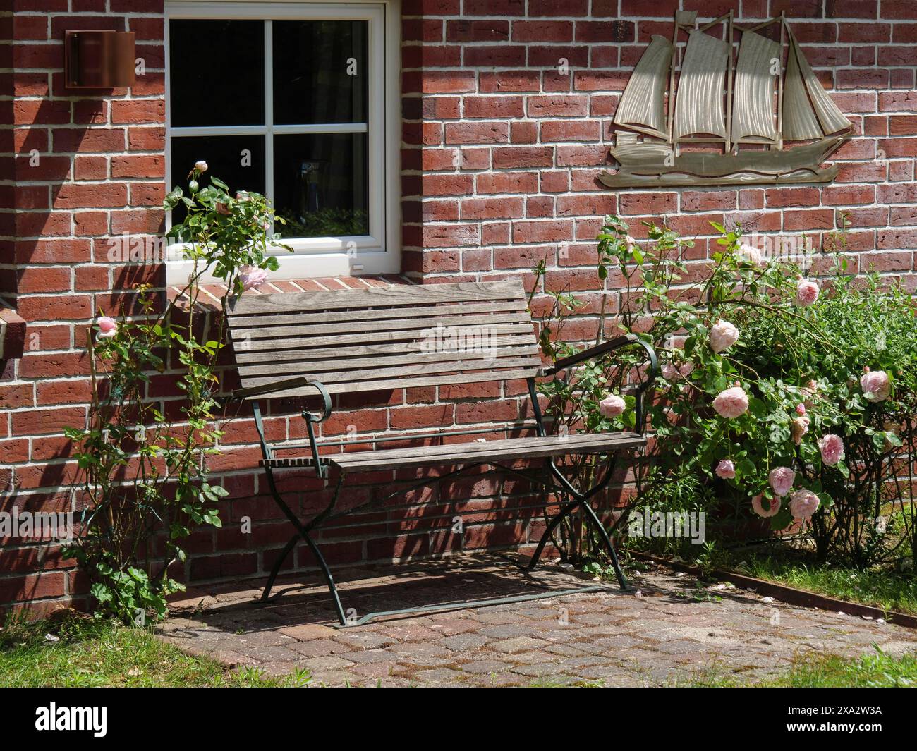 A wooden bench next to rose bushes in front of a red brick wall with a window, Baltrum Germany Stock Photo