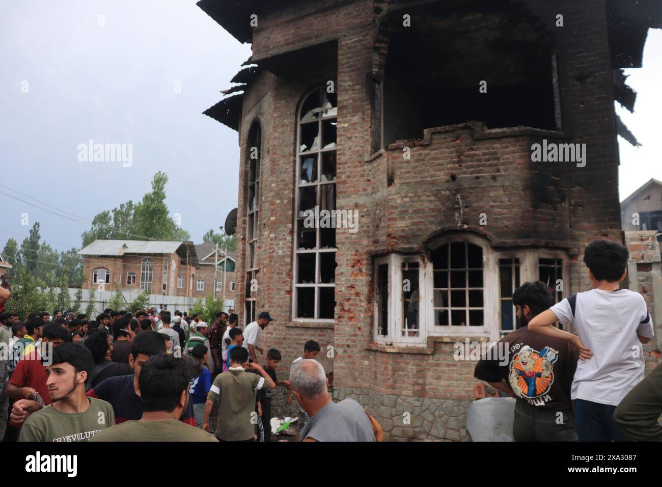 A crowd of people stands around a damaged and burnt multi-story brick building, observing the aftermath, Encounter, Pulwama, Jammu and Kashmir, India Stock Photo