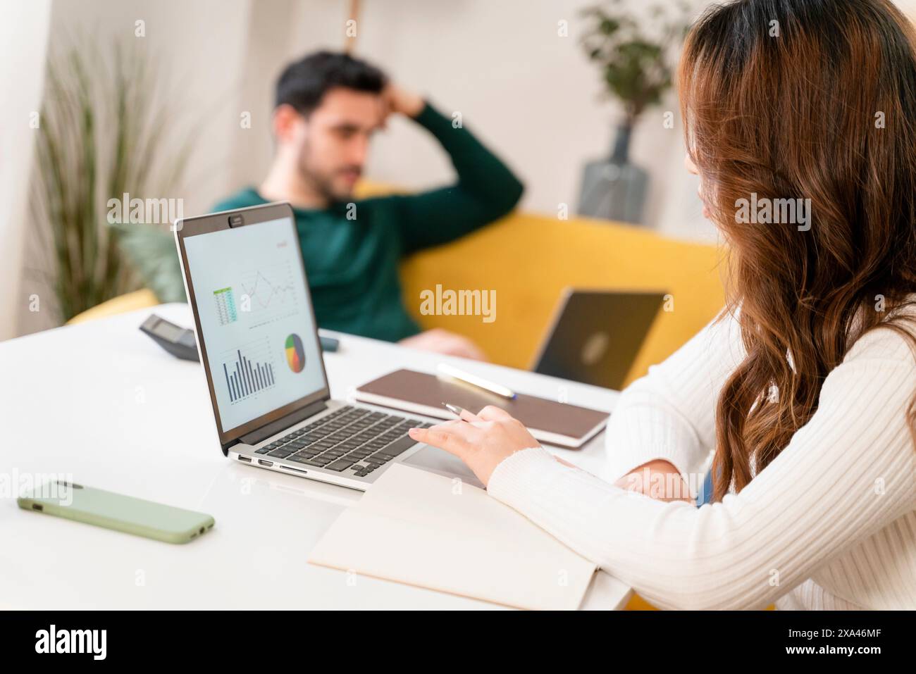 Woman working on laptop with charts as man relaxes in background Stock Photo